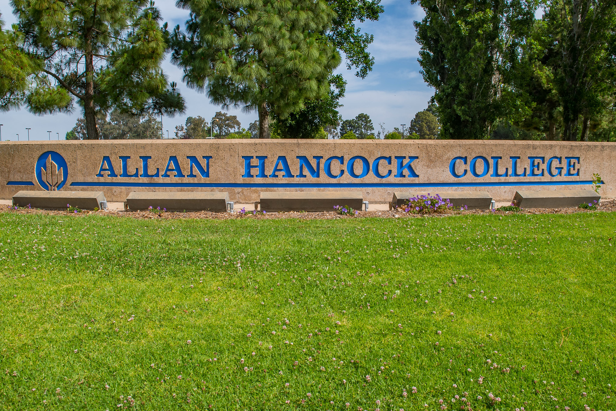 Entrance sign for Allan Hancock College featuring the college name and logo in blue lettering on a stone monument. The sign is surrounded by green lawn, flowers, and mature trees at the college’s Santa Maria campus.