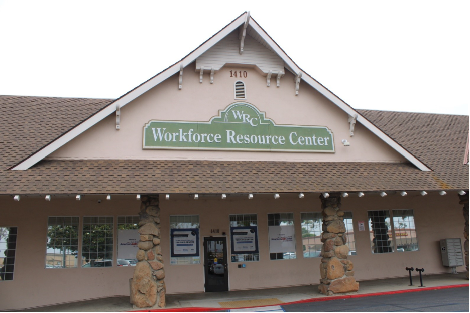 Photograph of a single-story building with a peaked roof labeled "Workforce Resource Center" on a green sign above entrance. Stone pillars flank the doorway, and windows display posters, indicating a facility for employment assistance and workforce services.