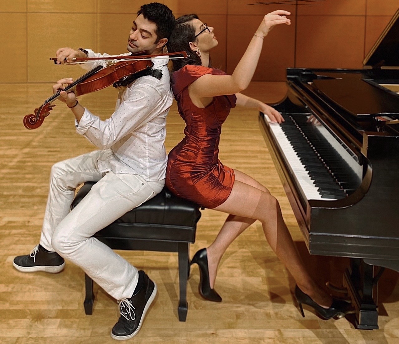 Allan Hancock College piano faculty member Dr. Adea Badivuku (right) and violist and composer Dr. Deniz Caglarcan (left) perform together on stage. Seated back-to-back on a piano bench, Badivuku plays a grand piano while Caglarcan plays the viola. The duo, winners of the Golden Classical Music Awards International Competition, perform in a concert hall setting