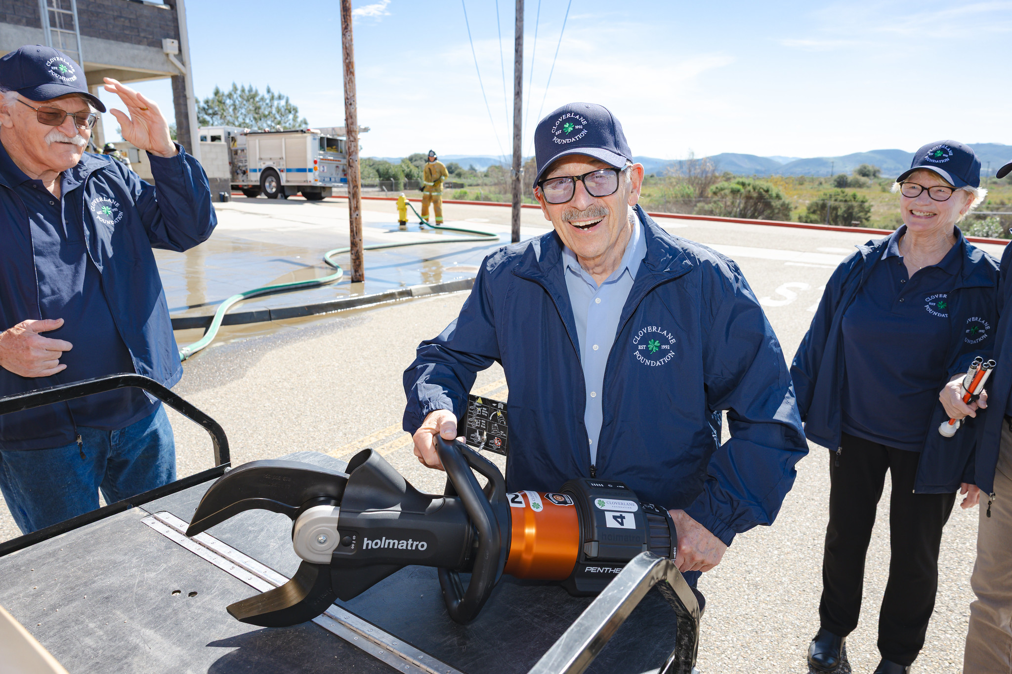Members of the Cloverlane Foundation stand outdoors holding a hydraulic rescue tool, commonly known as the ‘Jaws of Life,’ that they donated to the Allan Hancock College Fire Academy. The group wears matching navy jackets and hats with the foundation logo, while a fire engine and training area are visible in the background