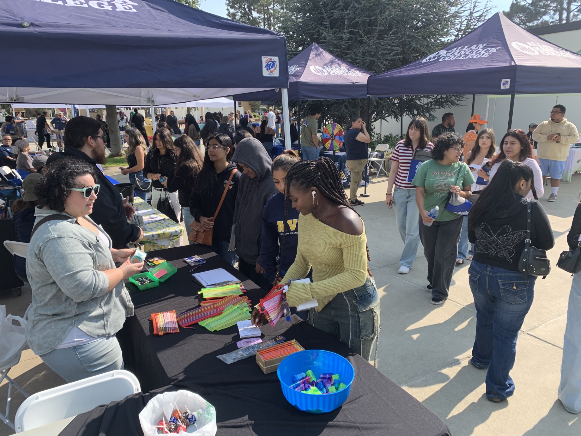 Students gather at Allan Hancock College’s Hancock Hello orientation event. Several students stand in line at an outdoor booth covered by a navy tent with the college logo, receiving colorful school supplies and information from staff members. Additional booths, canopies, and attendees are visible in the background on a sunny day.