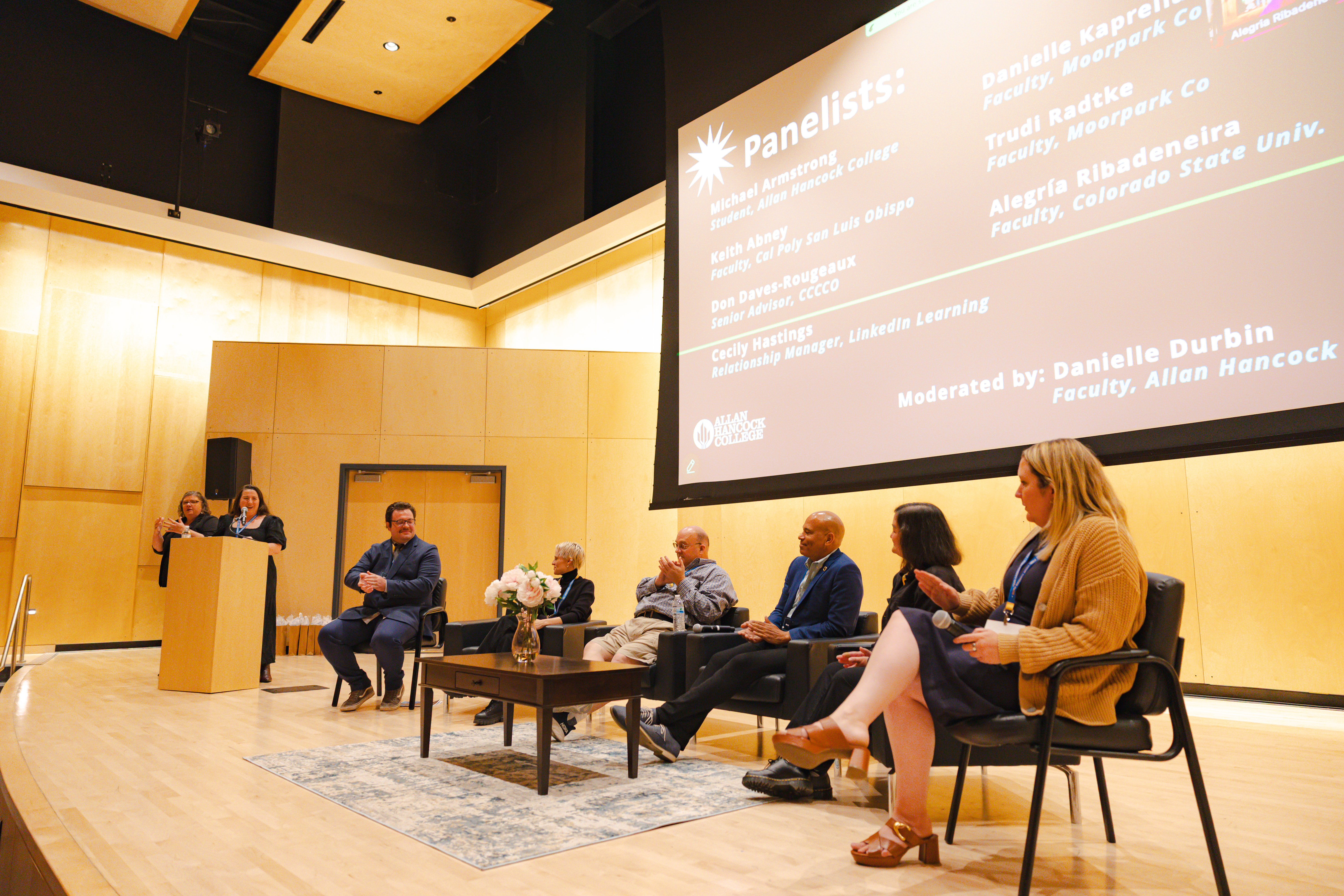 A panel of seven speakers sits on stage during Allan Hancock College’s first AI Summit. Two women stand at a podium speaking, while five panelists are seated in chairs, engaged in discussion. A large screen behind them displays the names of the panelists and moderator. The event is held in a bright, modern auditorium with wood-paneled walls.