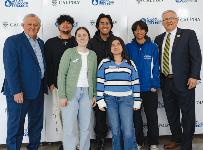 A group photo from a March 17 news conference features Allan Hancock College Superintendent/President Kevin G. Walthers, Ph.D. (left), and Cal Poly President Jeffrey D. Armstrong, Ph.D. (right), standing alongside five Allan Hancock College students. The students, dressed in casual and school-branded attire, are smiling and standing close together in front of a backdrop displaying the logos of Allan Hancock College and Cal Poly. The setting is indoors with bright lighting and a neutral-colored wall.