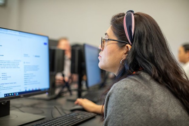 A woman wearing glasses and a pink headband focuses intently on a computer screen in a classroom or computer lab. Other people working at computers are visible in the blurred background.