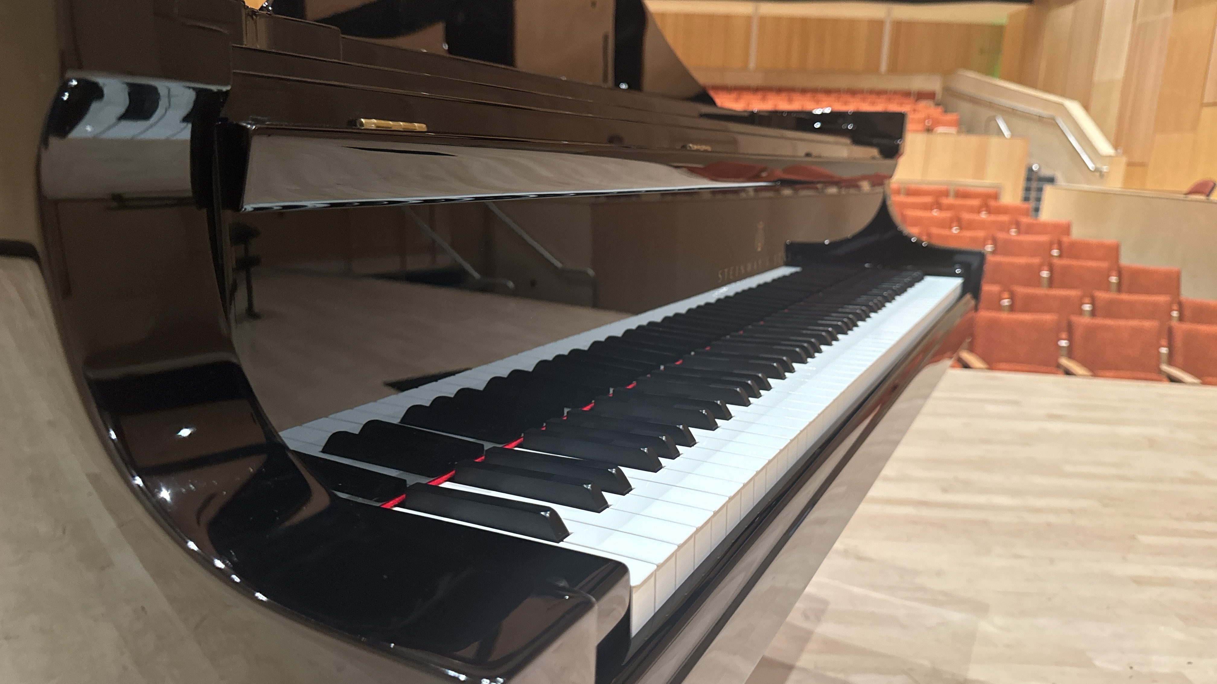 Close-up view of a Steinway & Sons grand piano on stage at Boyd Concert Hall at Allan Hancock College, with rows of empty orange seats and wood-paneled walls in the background.