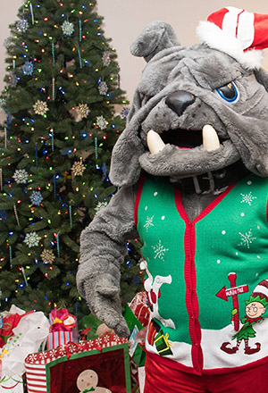 Spike mascot wearing a holiday sweater standing near a holiday tree