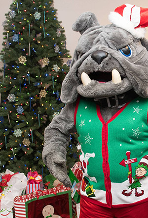 Spike mascot wearing a holiday sweater standing near a holiday tree