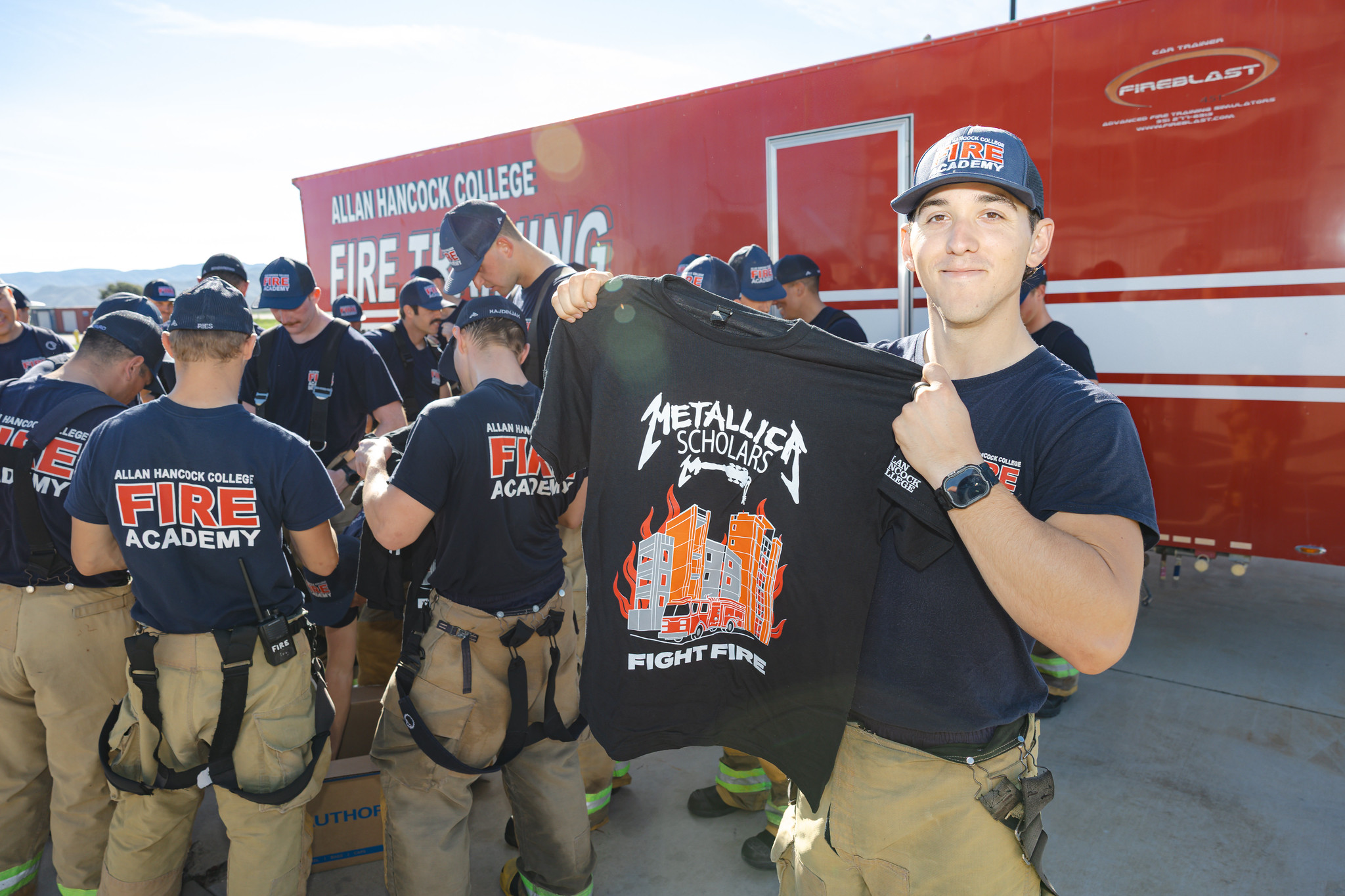 Metallica Scholars An Allan Hancock College Fire Academy cadet stands in front of the academy's fire training trailer, holding up a black Metallica Scholars 'Fight Fire' T-shirt. Around him, fellow cadets in Fire Academy uniforms gather and receive shirts before a training exercise, with the red training trailer visible in the background.